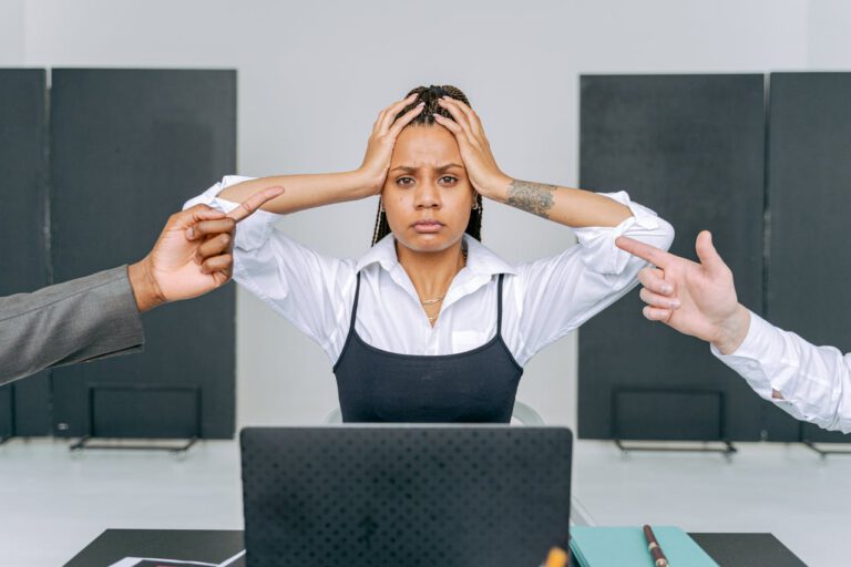 Woman Sitting in Front of Laptop with Head in Hands and Men Pointing at her
